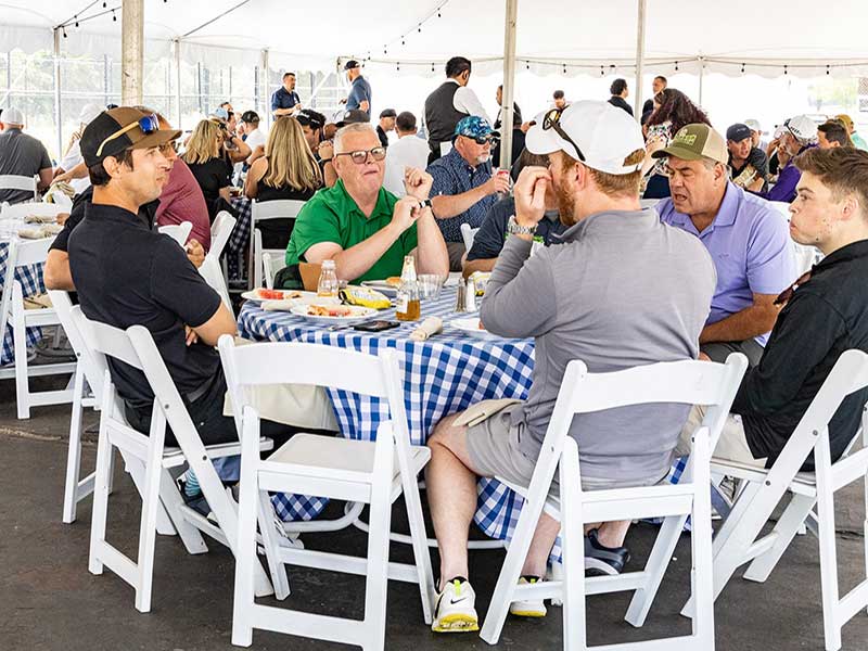 A group of seven men sit at a round table with a blue and white checkered tablecloth enjoy lunch at the IFPA Retail Golf event.