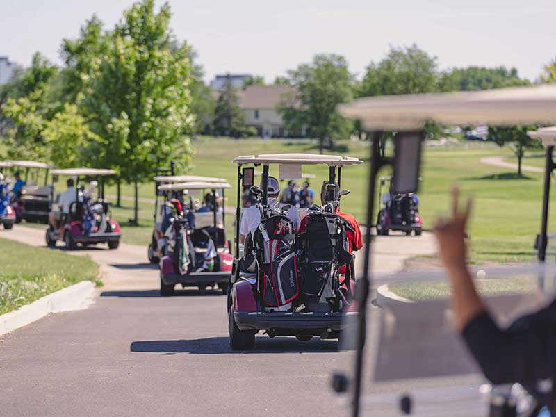 A view taken from behind of a  number of full golf cars proceed to the course.  The hands of the driver in the forefront holds his hand up with the peace sign.