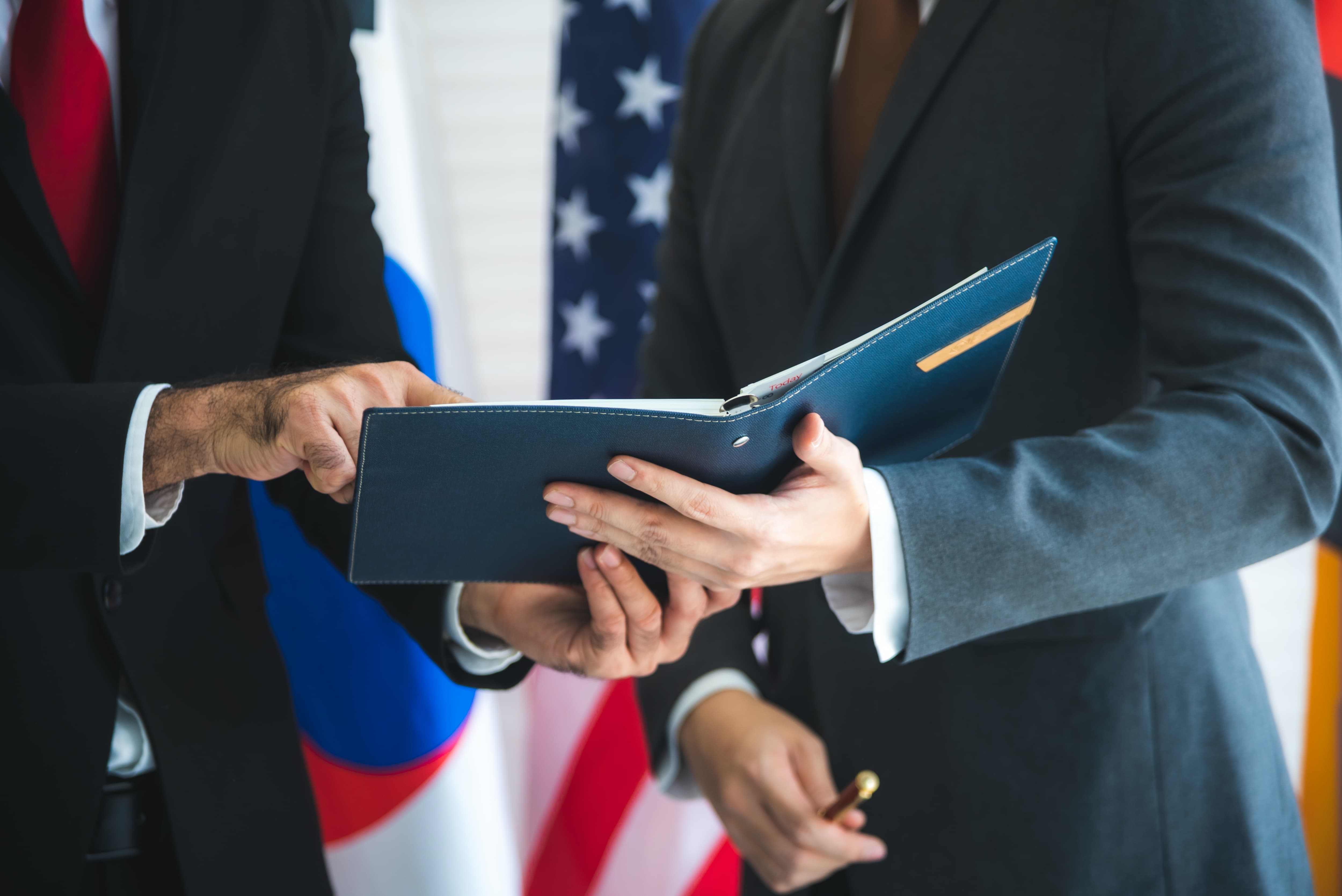 Two men in suits look over a proposal  while standing in front of flags.